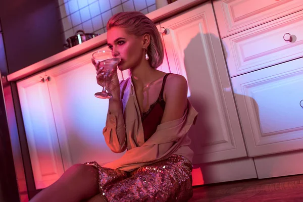 Bella donna elegante seduta vicino al bancone della cucina e bere cocktail in luce al neon — Foto stock
