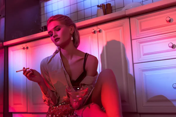 Schöne elegante Frau, die neben dem Küchentisch sitzt, Zigarette raucht und Cocktailglas im Neonlicht hält — Stockfoto