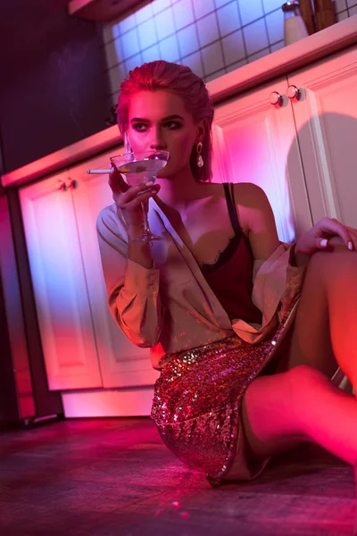Hermosa mujer elegante sentada cerca del mostrador de la cocina, fumar cigarrillo y celebración de copa de cóctel en luz de neón - foto de stock