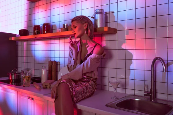 Selective focus of beautiful fashionable woman sitting on kitchen counter and smoking in neon light — Stock Photo