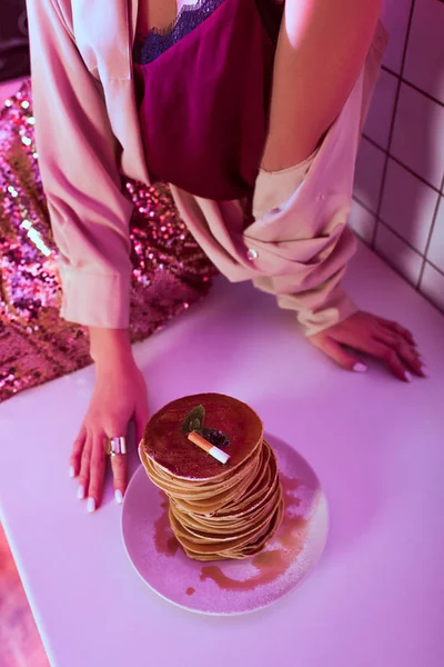 Cropped view of woman sitting on kitchen counter near plate of pancakes with cigarette ashes on top — Stock Photo