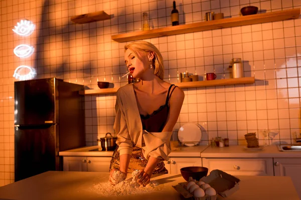 Elegante ama de casa con harina en el mostrador de cocina y fumar en la cocina con luz naranja - foto de stock