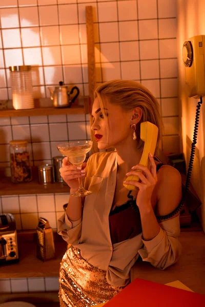 Beautiful elegant woman with cocktail talking on retro telephone in kitchen with orange light — Stock Photo