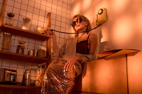 Elegant woman in sunglasses holding retro telephone in kitchen with orange light — Stock Photo