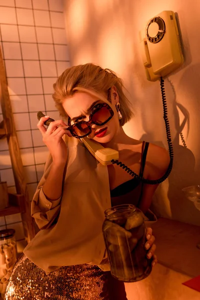 Beautiful elegant woman in sunglasses talking on retro telephone and holding jar with pickled cucumbers in kitchen with orange light — Stock Photo