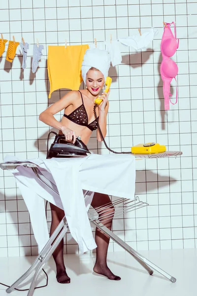 Hermosa sonriente ama de casa con toalla en la cabeza planchar ropa y hablando por teléfono retro en el baño - foto de stock