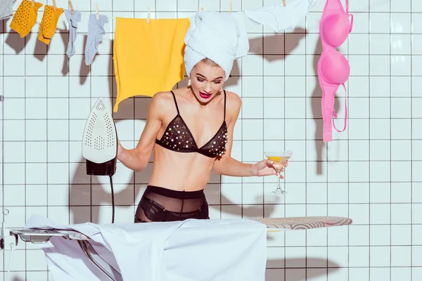 Удивленный домохозяйка в полотенце проведение коктейль стекло во время глажки в ванной комнате — стоковое фото