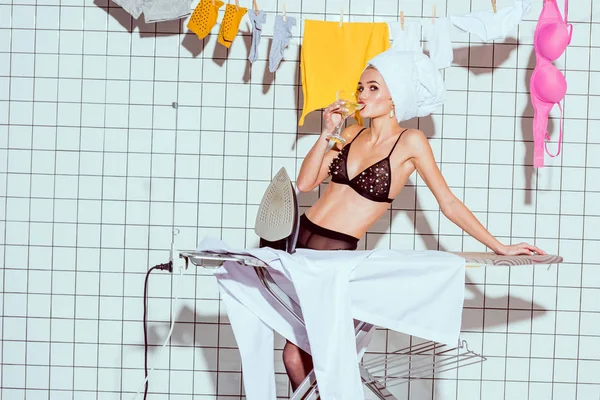 Beautiful housewife in towel drinking cocktail during ironing in bathroom — Stock Photo