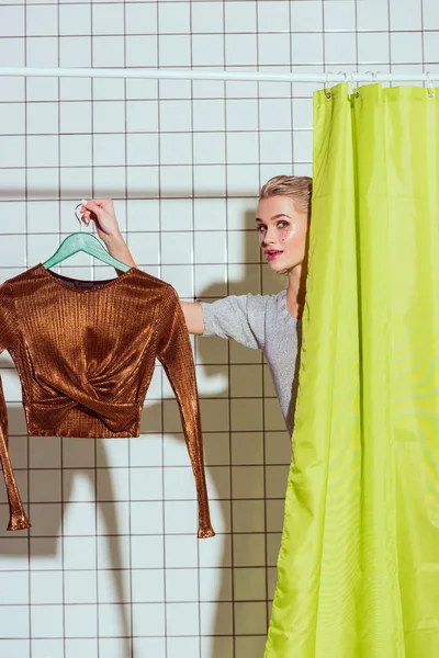 Woman holding bronze top on hanger and looking at camera in shower with green curtain — Stock Photo