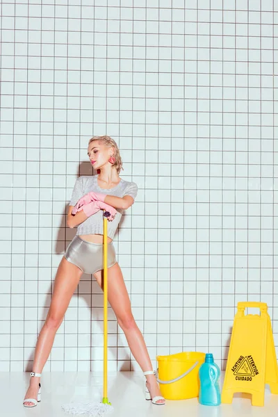 Jovem dona de casa segurando esfregona e posando com equipamentos de limpeza e azulejo branco no fundo — Fotografia de Stock