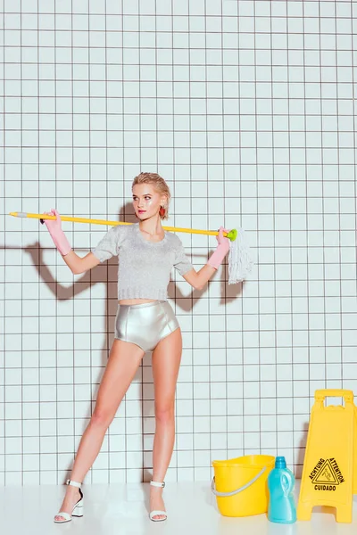 Bela dona de casa segurando esfregona e posando com equipamentos de limpeza e azulejo branco no fundo — Fotografia de Stock