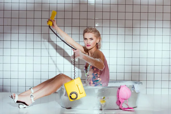 Mujer joven confundida en bañera de lavado con teléfono retro en el baño - foto de stock