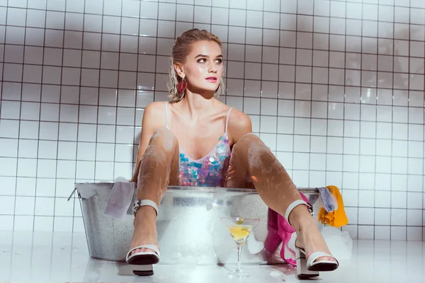 Beautiful young woman with cocktail glass sitting in wash tub in bathroom — Stock Photo