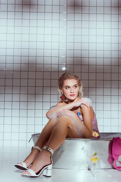 Bela jovem com coquetel de vidro sentado na banheira de lavagem no banheiro — Fotografia de Stock