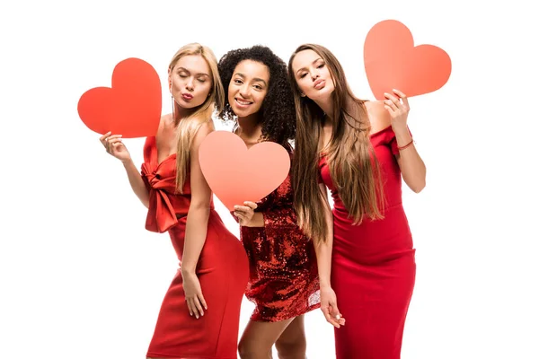 Hermosas chicas multiétnicas sosteniendo corazones de cartón, pucheros labios y posando aislados en blanco - foto de stock