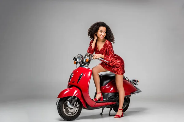 Beautiful smiling african american girl in red dress posing with motor scooter on grey background — Stock Photo