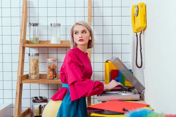 Beautiful housewife in colorful clothes listening music on vinyl record player and posing in kitchen — Stock Photo