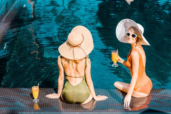 Vista traseira de pin up meninas em maiôs e chapéus sentados à beira da piscina com coquetéis laranja — Fotografia de Stock