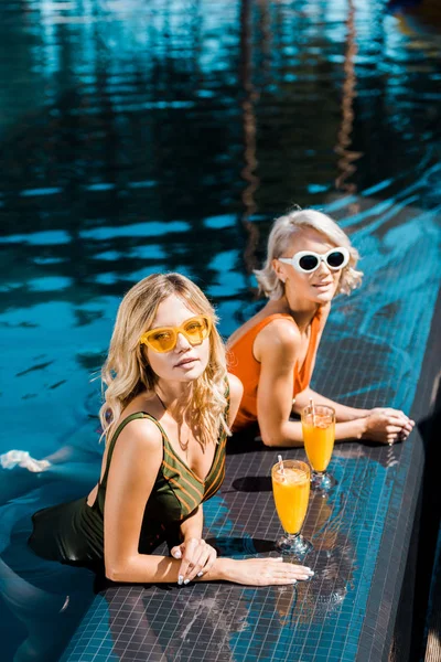 Rubia pin up niñas con vasos de jugo de naranja relajarse en la piscina - foto de stock