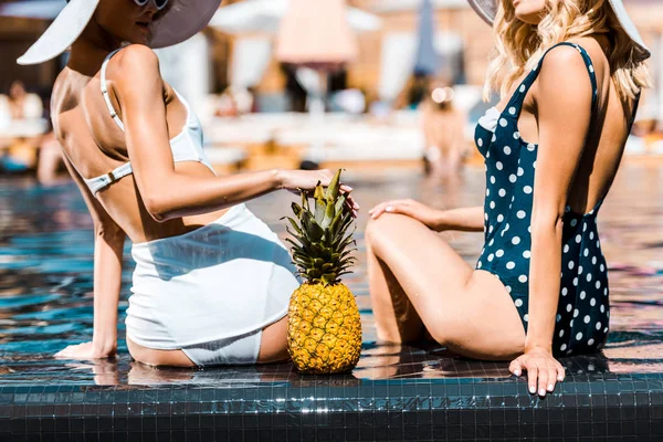 Cropped view of girls in pin up swimming suits sitting with pineapple at poolside — Stock Photo