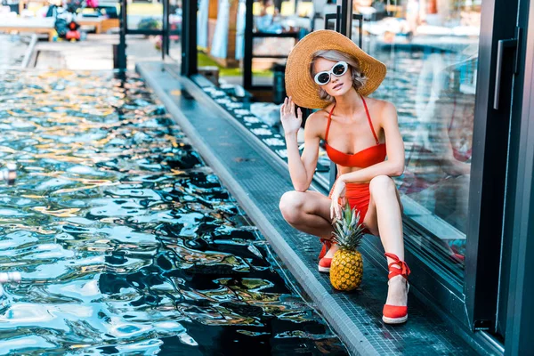 Atractiva mujer en traje de baño, gafas de sol y sombrero posando con piña cerca de la piscina - foto de stock