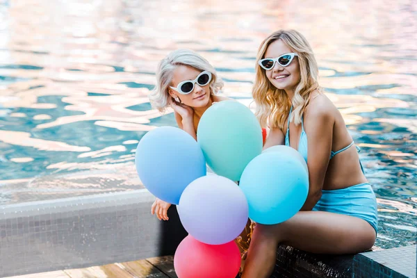 Happy pin up girls in swimwear holding balloons while sitting at poolside — Stock Photo