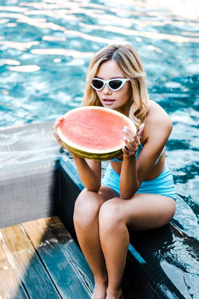 Привлекательная блондинка в купальнике и солнцезащитных очках, позирующая с арбузом у бассейна — стоковое фото