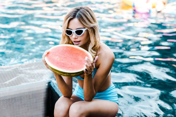 Blonde girl in swimsuit and sunglasses posing with watermelon at poolside — Stock Photo