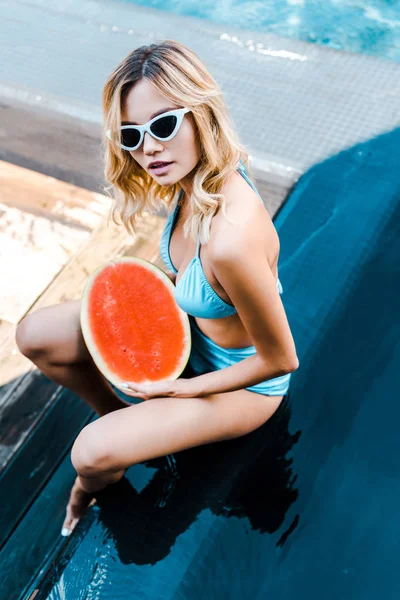 Blonde young woman in swimsuit and sunglasses posing with juicy watermelon at poolside — Stock Photo