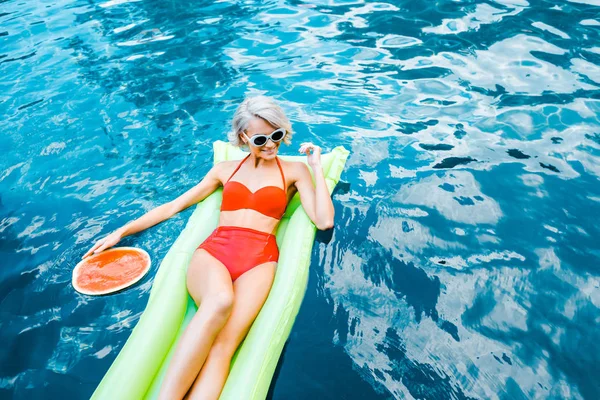 Happy pin up girl in swimsuit relaxing on green inflatable mattress in swimming pool with watermelon — Stock Photo