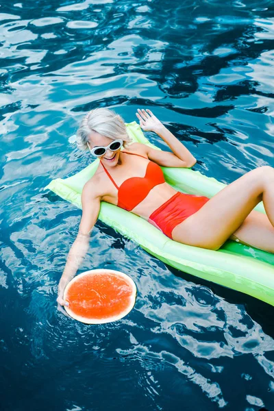 Lächelndes Mädchen im Badeanzug entspannt auf grüner aufblasbarer Matratze im Schwimmbad mit Wassermelone — Stockfoto