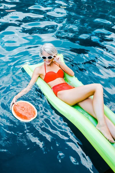 Smiling blonde woman in swimsuit relaxing on green inflatable mattress in swimming pool with watermelon — Stock Photo