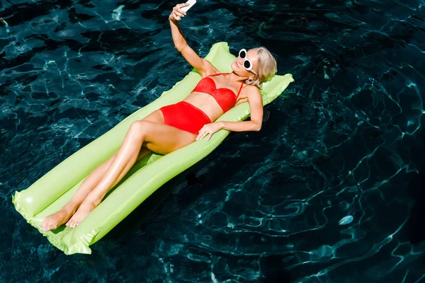 Красивая девушка в купальнике делает селфи на смартфоне, лежа на зеленом надувном матрасе в бассейне — стоковое фото