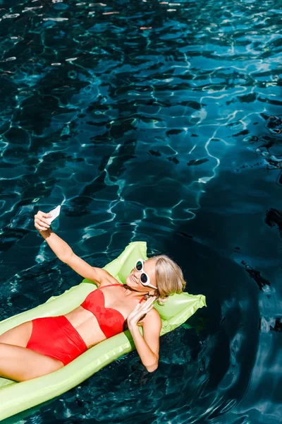 Atractiva chica en traje de baño saludando y tomando selfie mientras se relaja en el colchón inflable verde en la piscina - foto de stock