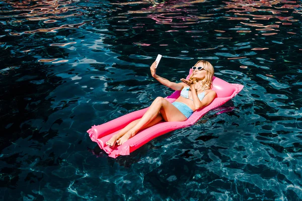 Menina bonita em maiô tomando selfie enquanto relaxa no colchão inflável rosa na piscina — Fotografia de Stock