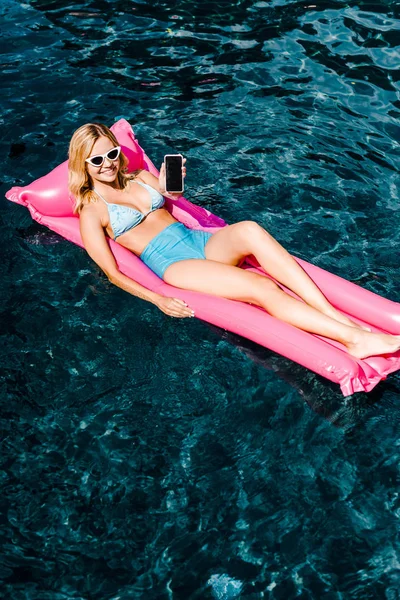 Attractive girl in swimsuit lying on pink inflatable mattress in swimming pool and showing smartphone — Stock Photo