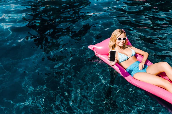 Beautiful girl in swimsuit lying on pink inflatable mattress in swimming pool and showing smartphone with blank screen — Stock Photo