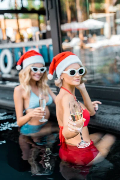 Selective focus of girls in swimsuits and santa hats holding champagne glasses and relaxing in swimming pool — Stock Photo