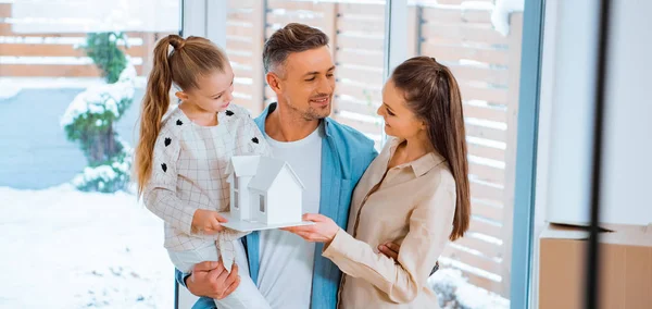Handsome father holding in arms cheerful daughter while standing near wife with house model in hands — Stock Photo