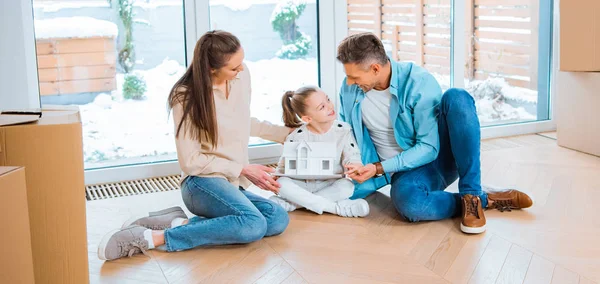 Pai alegre olhando para a filha com modelo de casa em mãos perto da esposa enquanto sentado no chão em nova casa — Fotografia de Stock