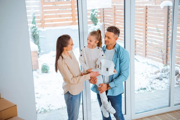 Feliz padre sosteniendo en brazos hija con modelo de casa en las manos mientras mira a la esposa - foto de stock