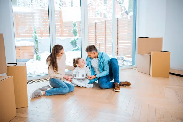 Padre feliz mirando a la hija con modelo de casa en las manos cerca de la esposa mientras está sentado en el suelo en un nuevo hogar - foto de stock