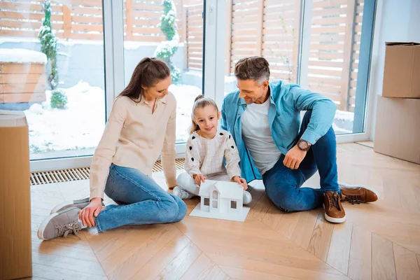 Happy husband and wife looking at cute daughter with house model in hands while sitting on floor in new home — Stock Photo