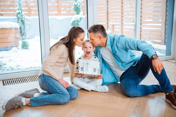 Feliz marido y esposa besando linda hija con modelo de casa en las manos mientras está sentado en el suelo en un nuevo hogar - foto de stock