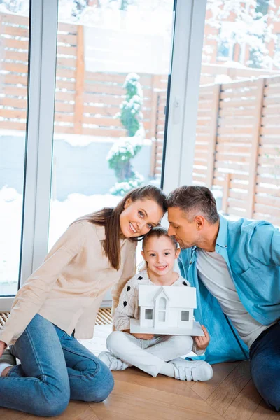 Glücklicher Vater küsst süße Tochter mit Hausmodell in den Händen, während er auf dem Boden neben Frau sitzt — Stockfoto