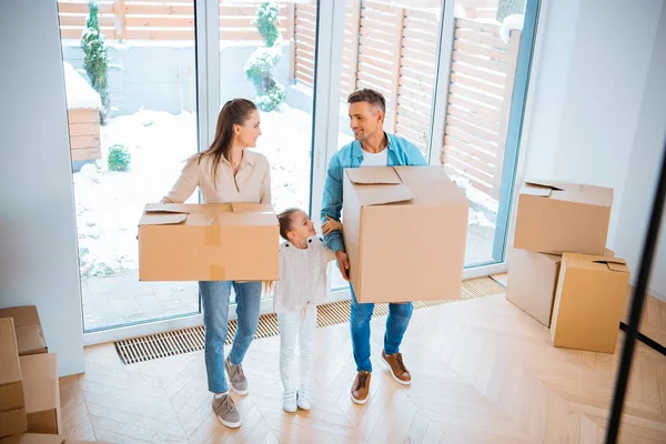 Kid standing between cheerful mother and father looking at each other while holding boxes in new home — Stock Photo