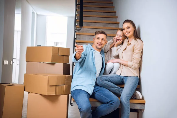 Cheerful husband pointing with finger and smiling while sitting on stairs near wife and daughter in new home — Stock Photo