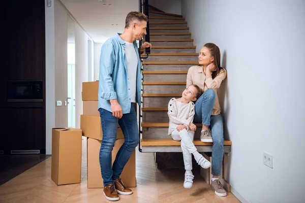 Hombre guapo mirando a la hija y la esposa sentado en las escaleras en el nuevo hogar - foto de stock