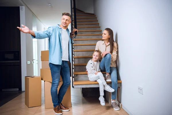 Hombre guapo mostrando algo cerca linda hija y esposa sentado en escaleras en nuevo hogar - foto de stock