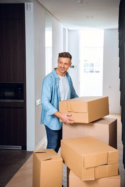 Guapo hombre sosteniendo caja en las manos y sonriendo en nuevo hogar - foto de stock